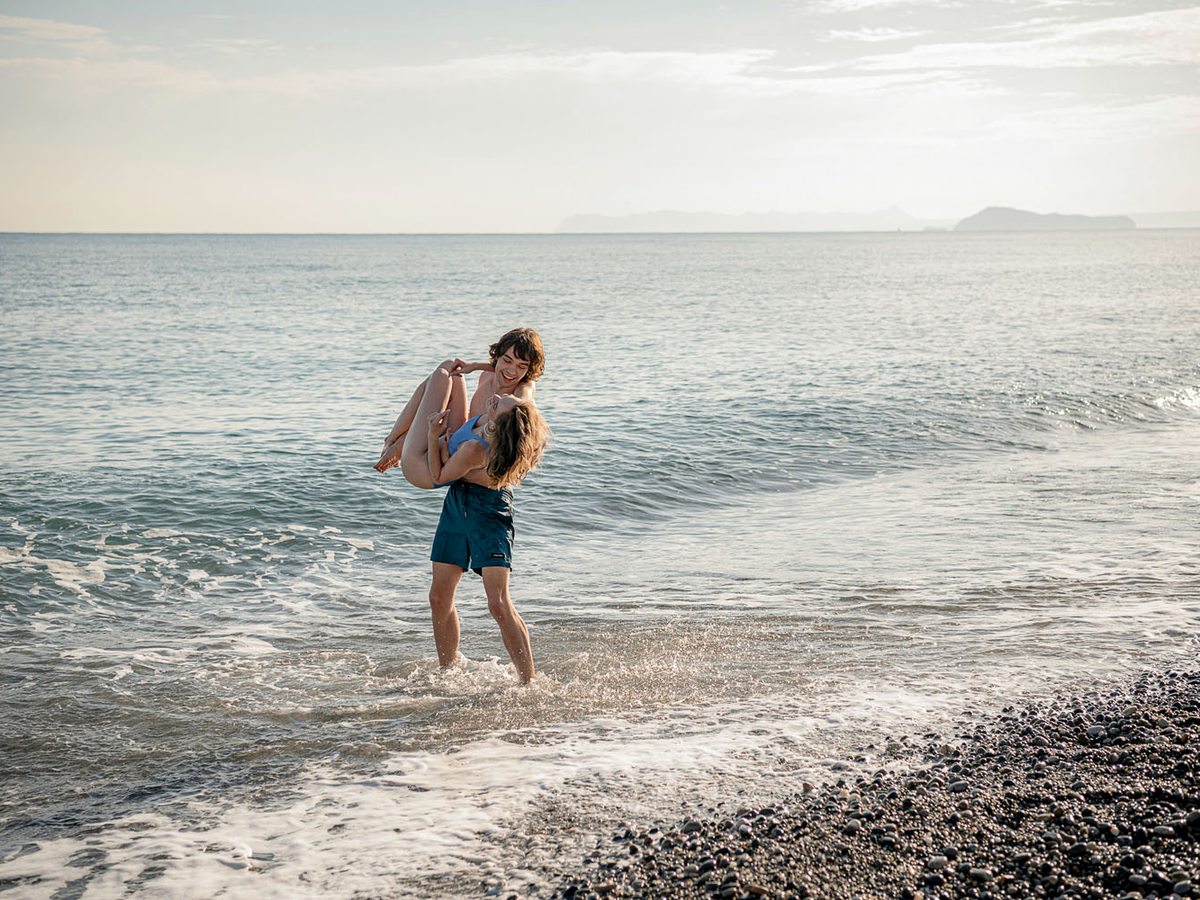 An adult carrying a young child into calm shallow water off the pebble beach, an island low on the horizon.