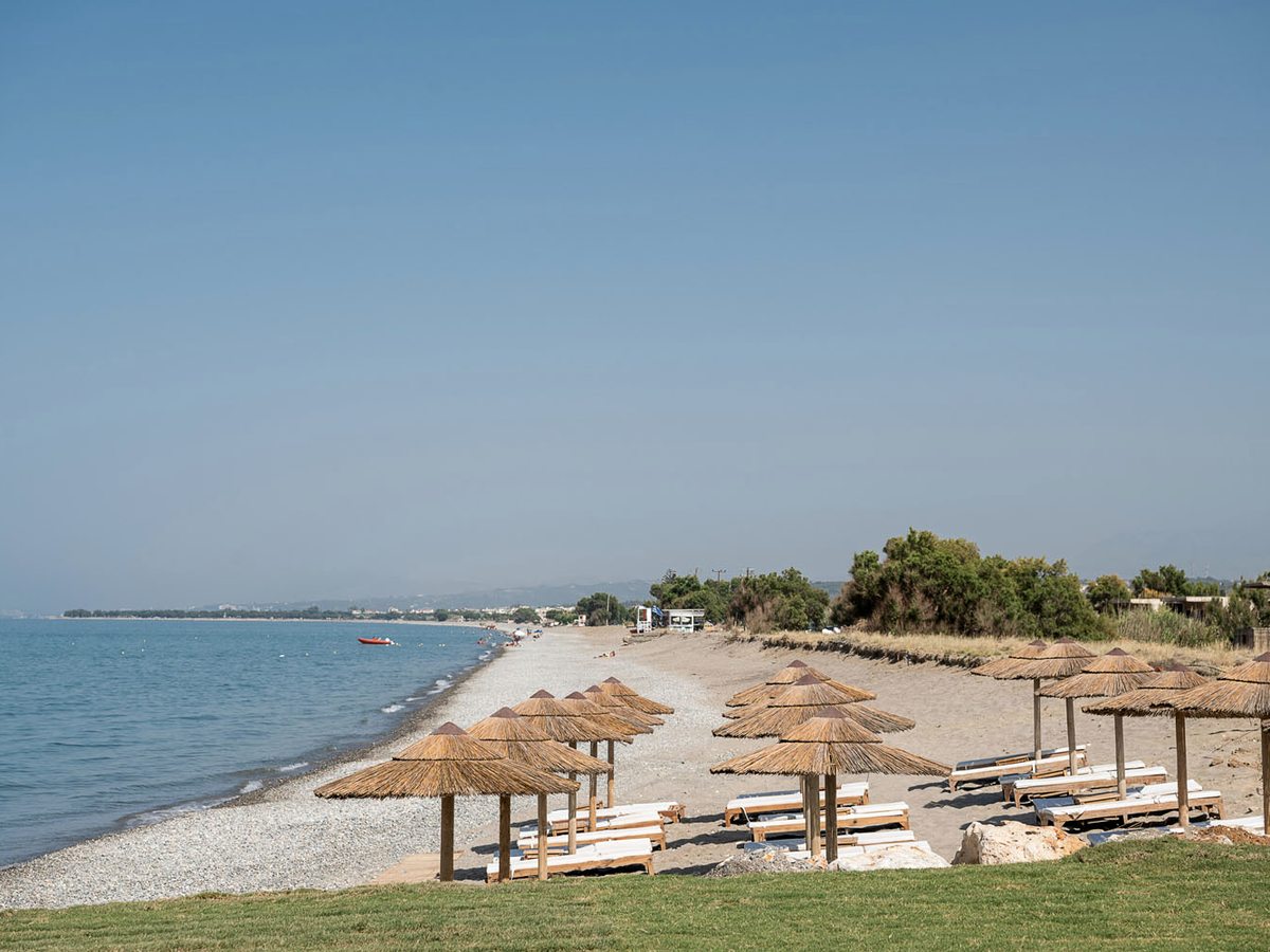 A wide daytime view along Kolymvari beach, straw parasols and wooden loungers in the foreground and the pebble shore curving away past a grassy bank towards distant hills.