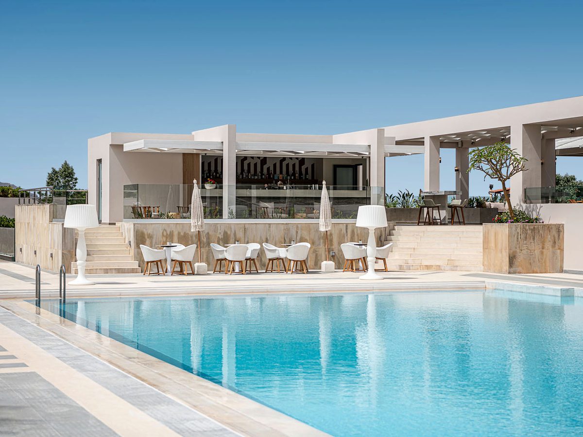A covered stone-counter bar on a terrace above a pool, with cafe tables, chairs and tall white floor lamps.