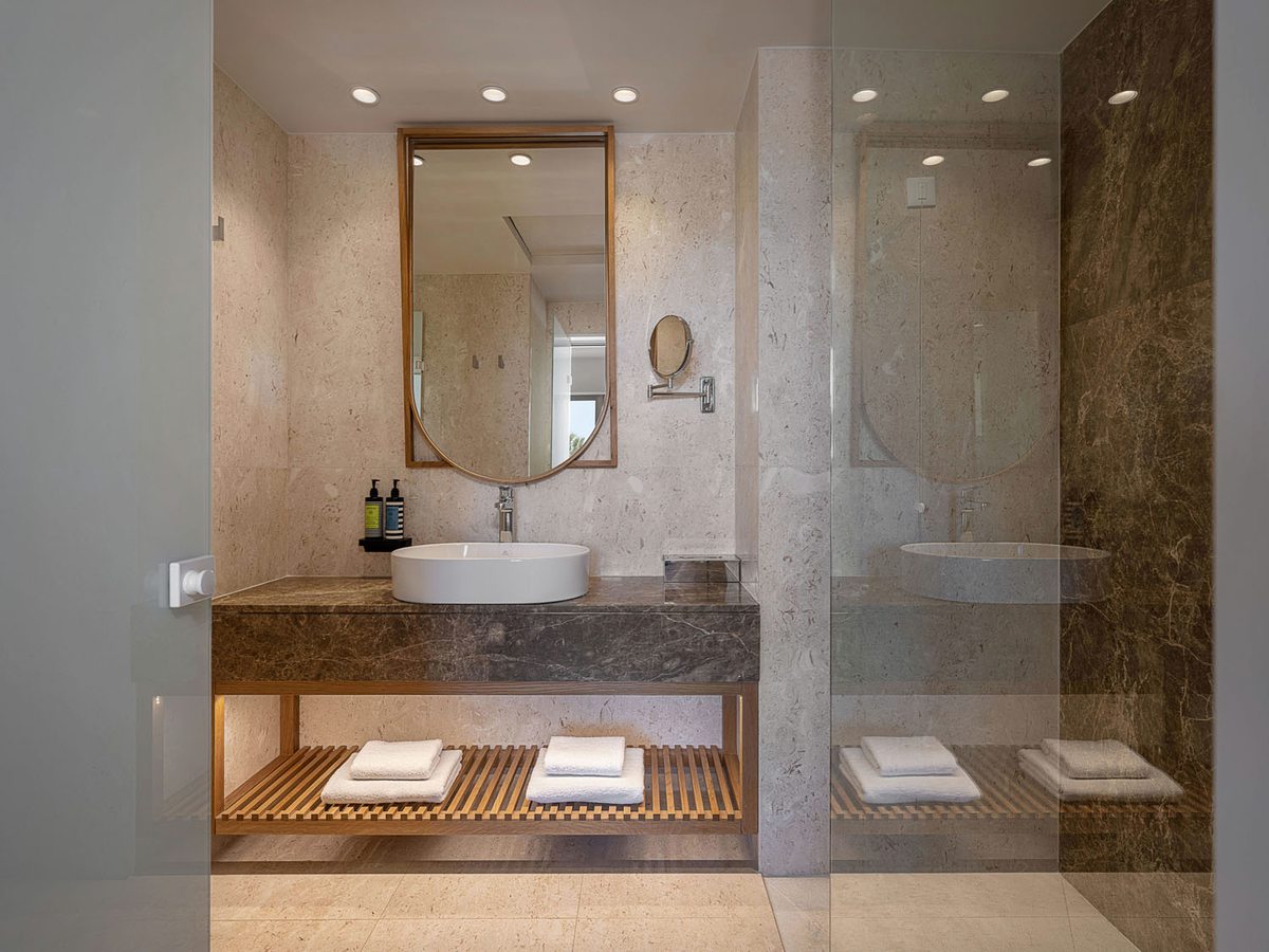 A marble bathroom with a round basin, a lit mirror and a walk-in shower behind a glass screen.