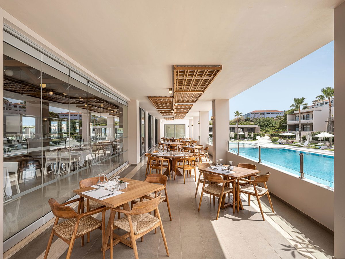 A covered restaurant terrace of wooden tables and woven chairs beside a pool, glazed along one side.
