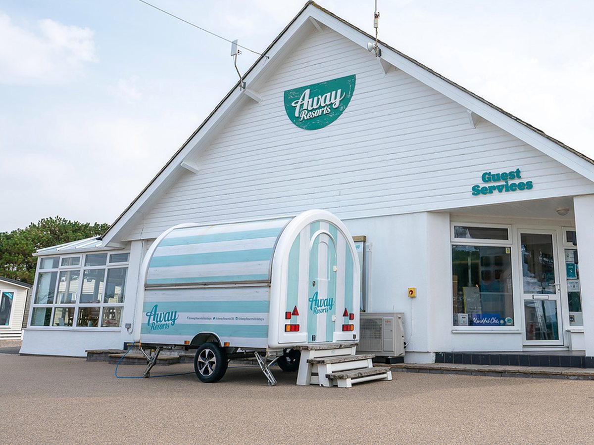 A white clapboard reception building with turquoise Away Resorts signage and a branded caravan parked outside.