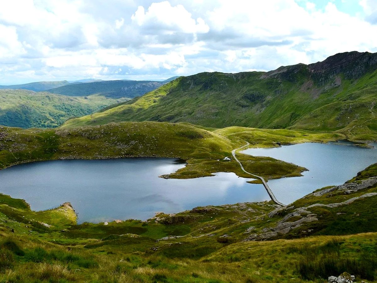 A still mountain lake ringed by green and rocky hills under a cloudy sky.