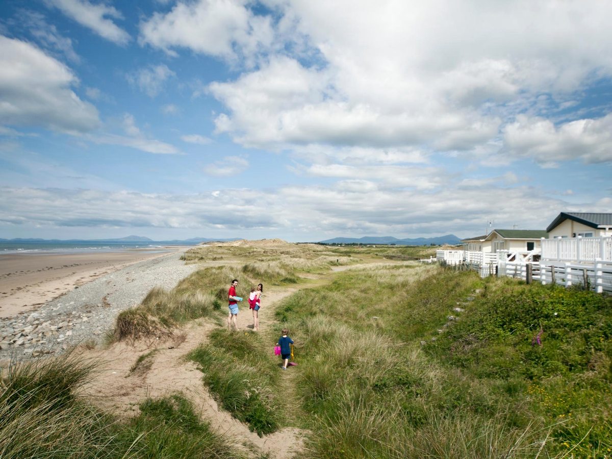 A sandy path running through marram-grass dunes towards a wide beach, with a family walking down and caravans to the side.