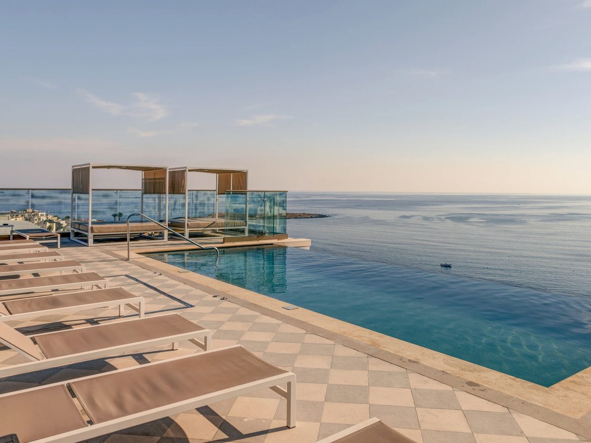 A pool terrace with cushioned cabana daybeds and tan sun loungers on a checkerboard deck, beside an infinity-edge pool that meets the calm sea of the bay.