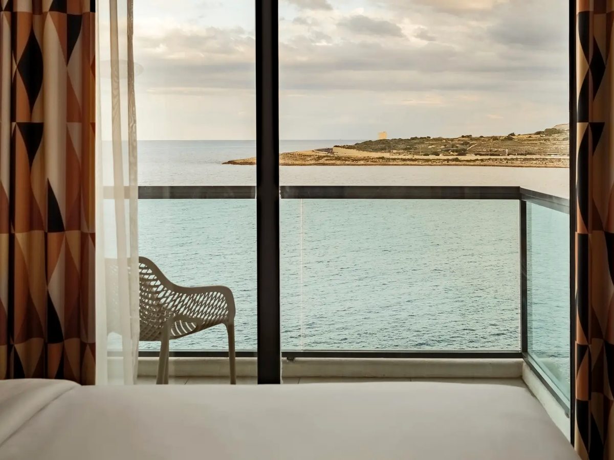 A hotel bedroom with patterned curtains framing a floor-to-ceiling window and a woven balcony chair on a glass-railed balcony, looking out over calm sea to a low rocky headland.