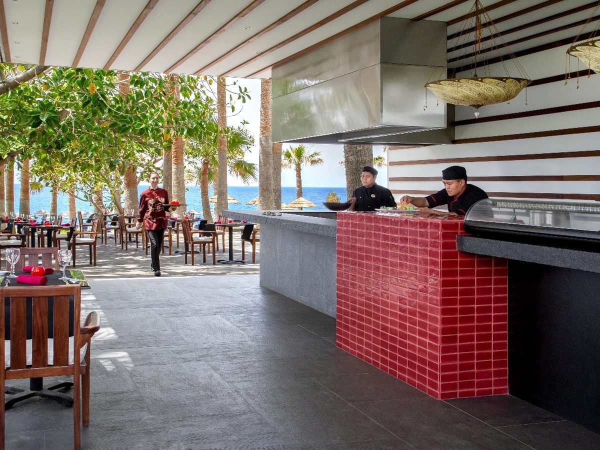 A red-tiled open kitchen counter with chefs cooking, beside a covered terrace looking out to sea.
