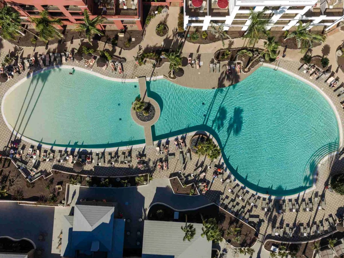 An aerial view looking down on a curved swimming pool ringed by sun loungers, palms and low-rise accommodation blocks.