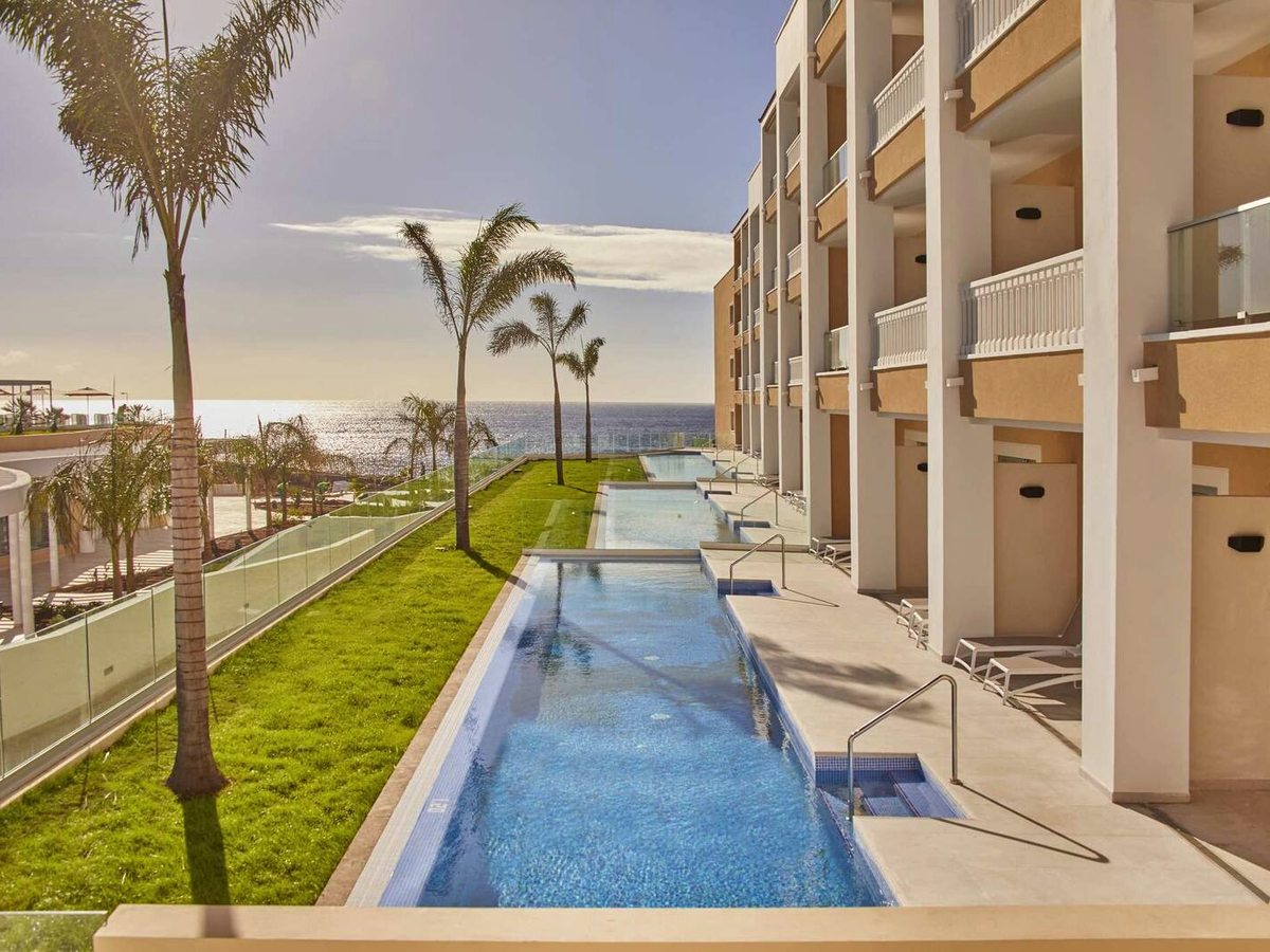 A long narrow swim-up pool running between a grassy strip with palms and a wing of ground-floor rooms, the sea beyond.