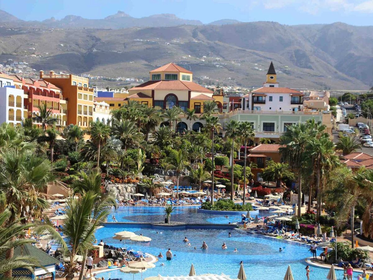 Busy outdoor pool among palms and landscaped grounds, with a domed resort building and Tenerife's volcanic mountains rising behind.