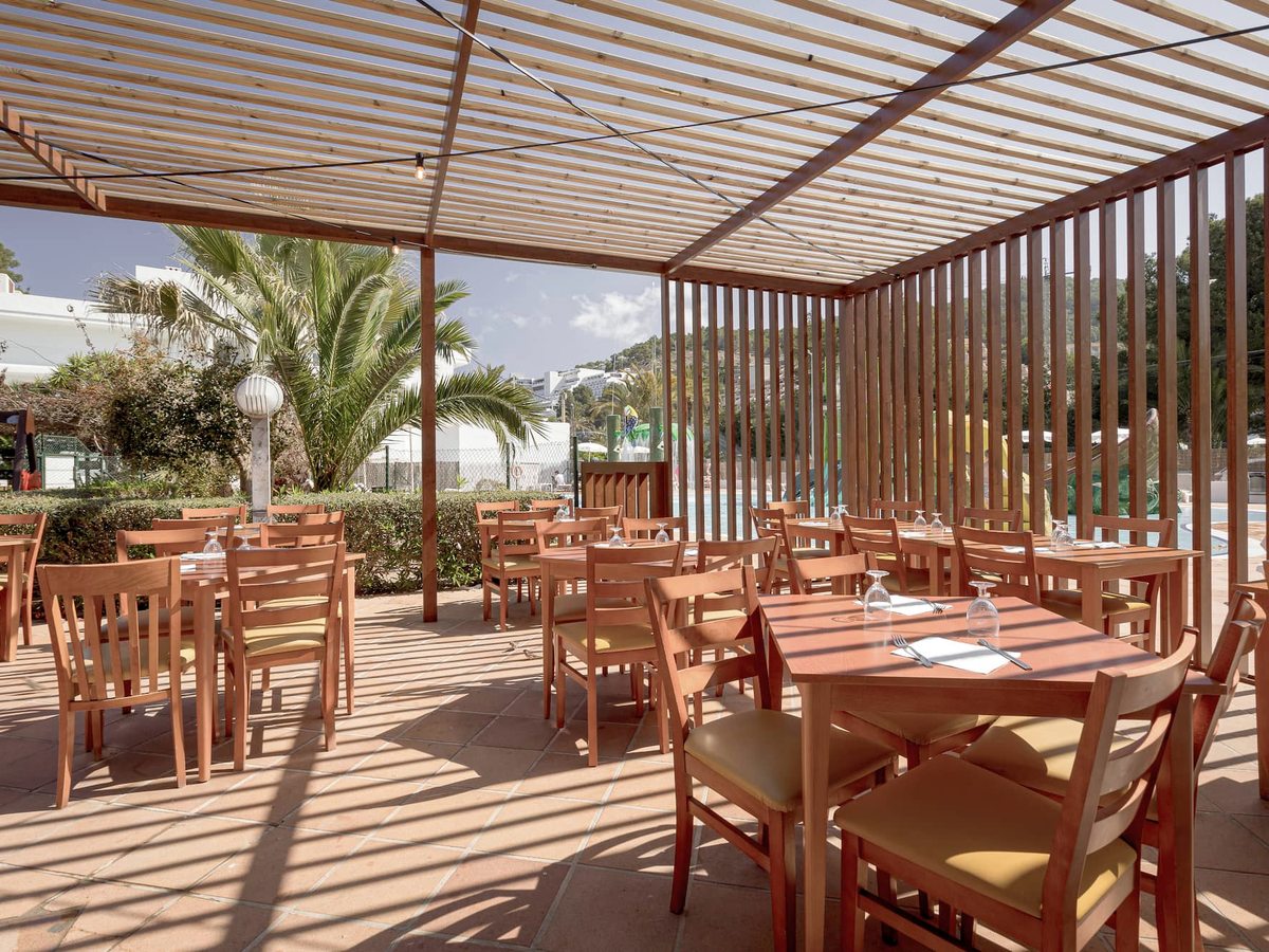 Covered outdoor dining terrace with wooden tables and chairs under a slatted timber roof, open to greenery on one side.