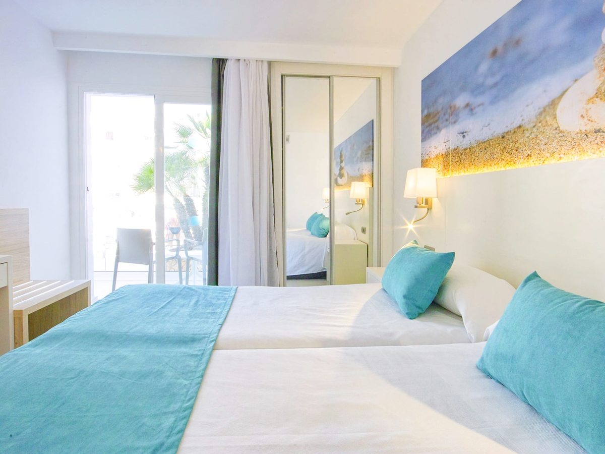 Bedroom with twin beds pushed together under turquoise pillows and a beach-themed headboard, with a wall mirror and sheer curtains.