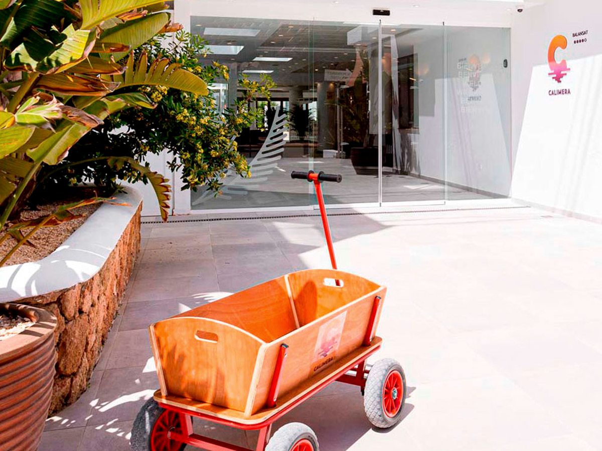Covered white entrance walkway lined with low planting, leading to the glass reception doors, with a small orange hand cart to one side.
