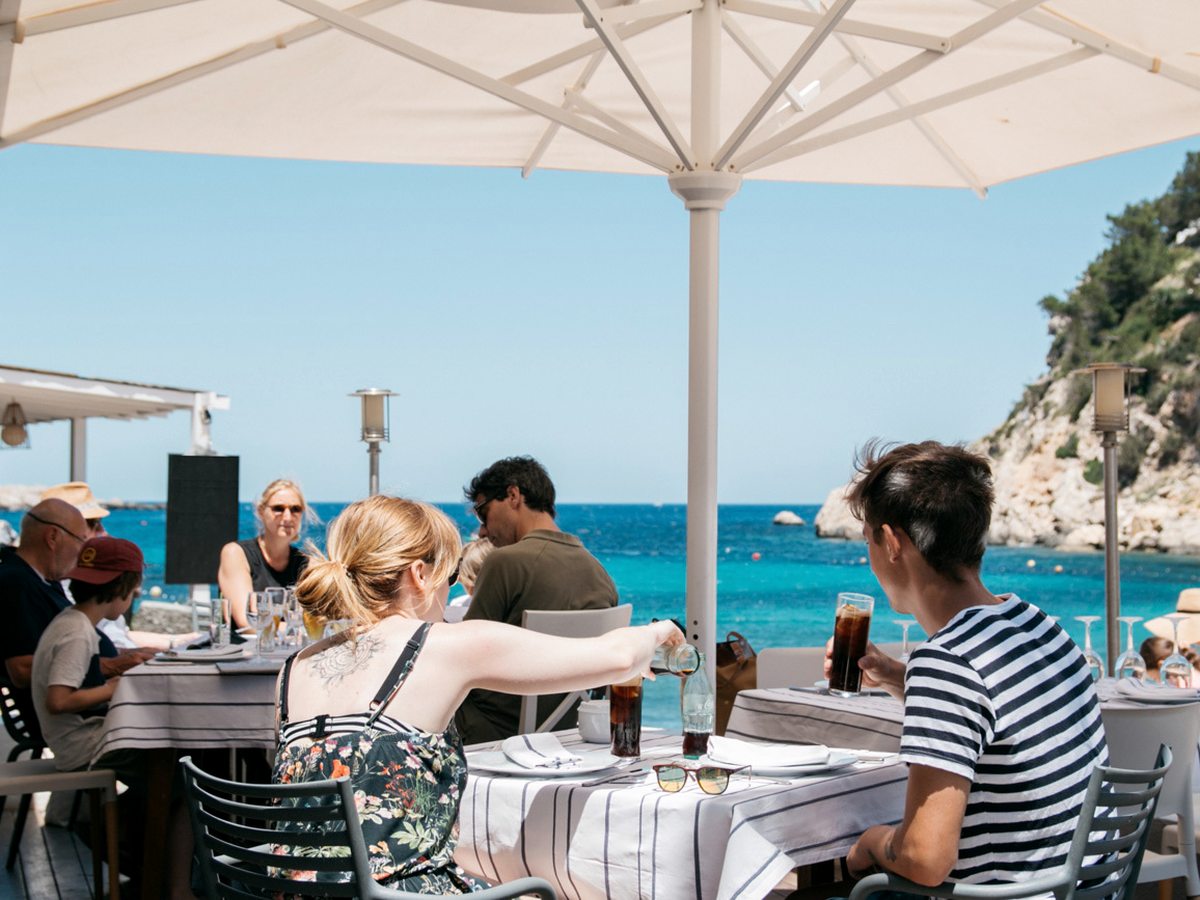 White-clothed tables under a large parasol on a seafront terrace, guests dining above the turquoise cove and cliffs.