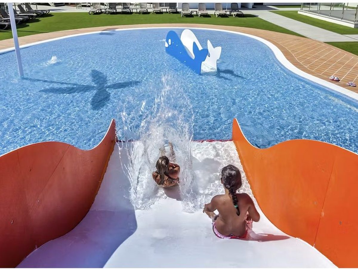 Two children sliding down a curved white-and-orange slide into a shallow kidney-shaped pool with a blue whale water feature, sun loungers on the grass behind.