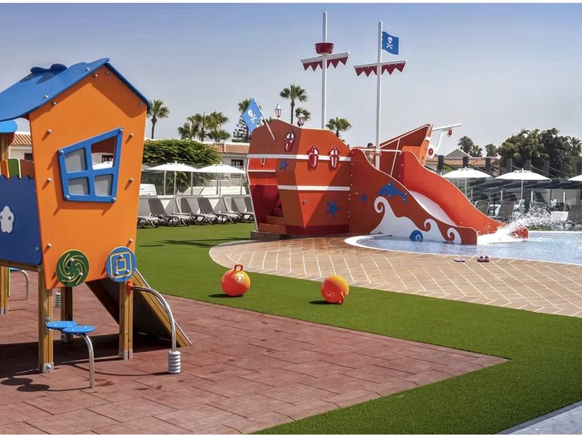 A children's play area on artificial grass with a wooden climbing frame and playhouse and, beyond, an orange pirate ship with a slide into a shallow blue pool.
