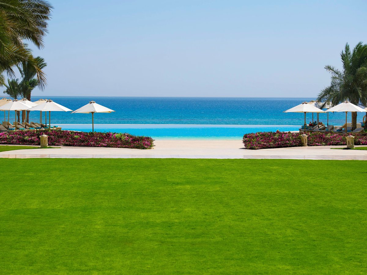 A wide manicured lawn edged with flower borders leading to white beach parasols, the sandy shore and the turquoise sea.