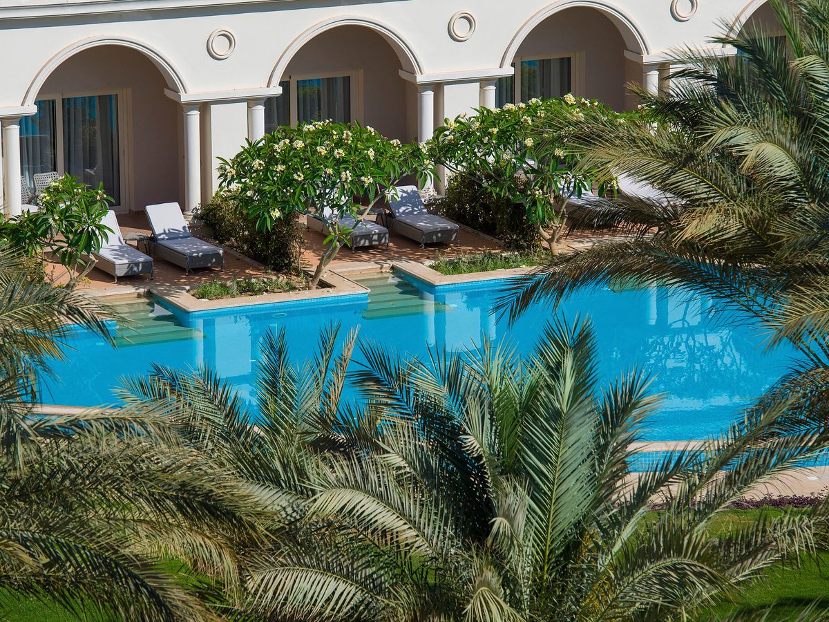 A lagoon-style pool running past arched ground-floor room terraces, with sun loungers, palms and garden planting.