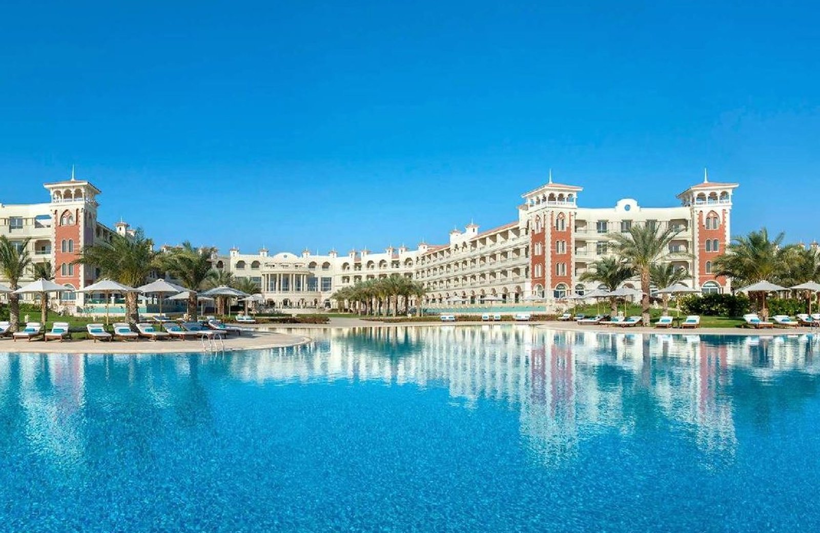 Baron Palace Sahl Hasheesh: the resort's turreted white and terracotta facade reflected in the large main pool under a clear blue sky, with sun loungers and parasols along the poolside.