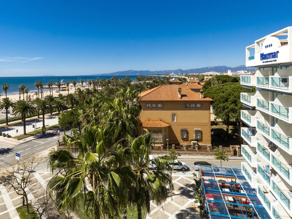 An aerial with the sandy beach and sea on the left, a palm-lined promenade, and the Blaumar hotel on the right