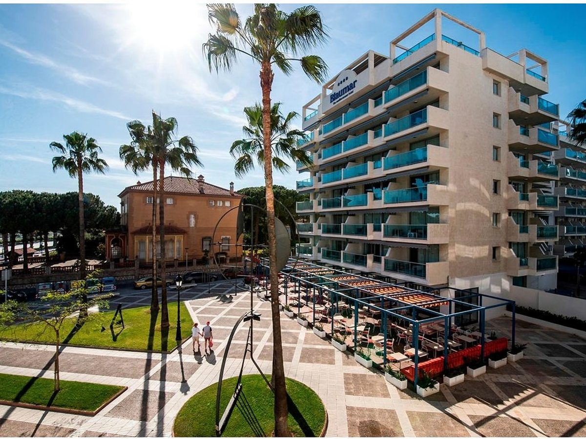 The white Blaumar hotel tower with glass balconies beside tall palms and a tiled promenade plaza, a restaurant terrace at its base