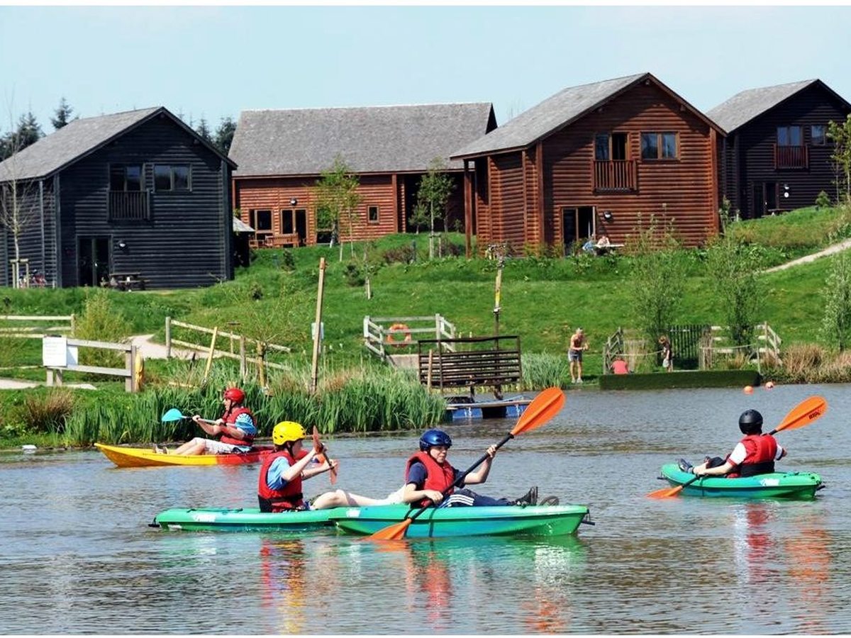 Families kayaking in green kayaks on a calm lake, with timber lodges on the grassy bank behind.