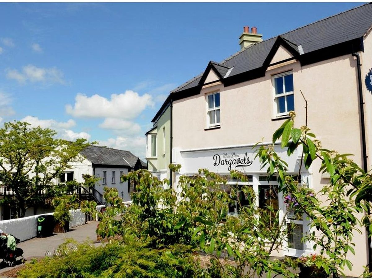 A cream-rendered Village building with a shopfront and slate roof, a planted path and a parent pushing a pram, under a blue sky.