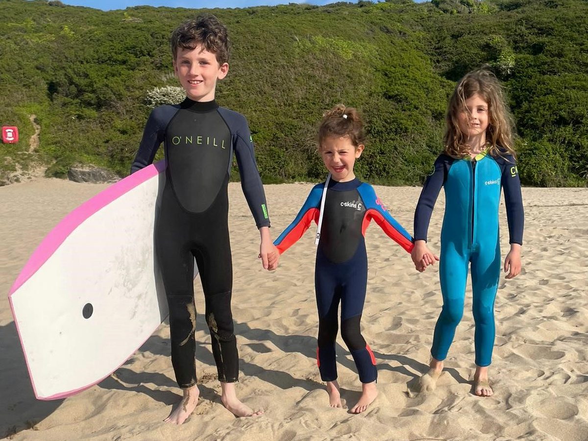 Three children in wetsuits, one holding a bodyboard, standing on a sandy Cornish beach backed by green cliffs.