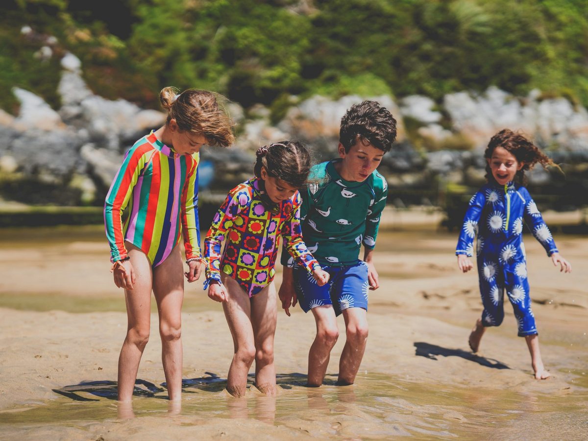 Children in swimwear and wetsuits paddling out of the shallow sea onto a sandy beach with a rocky shoreline behind.