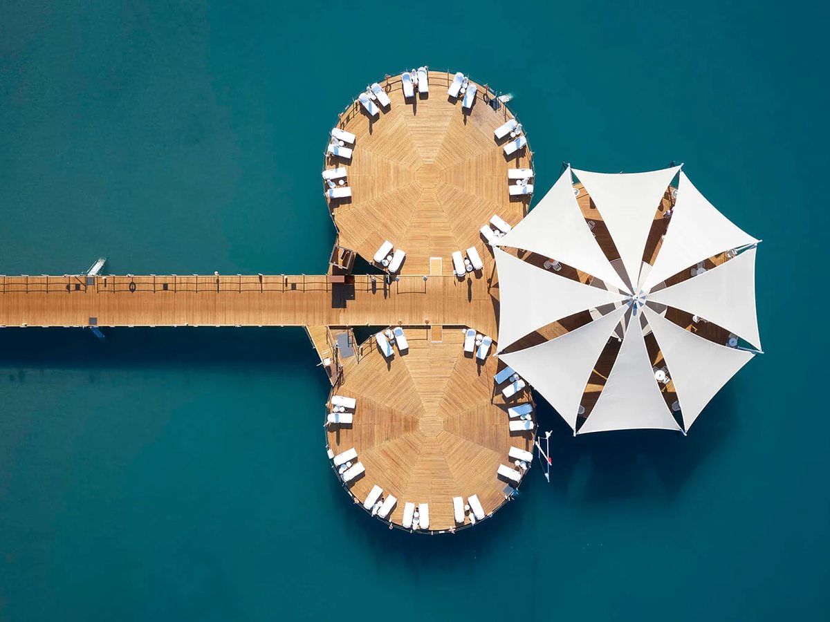 Aerial view of a circular timber sun deck at the end of a pier, ringed with loungers and a large white parasol over turquoise sea.