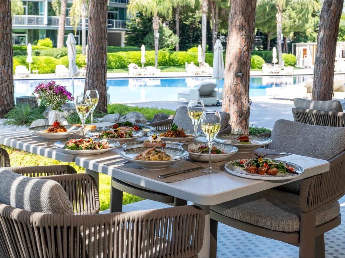 A covered poolside terrace with a laid dining table of plated dishes, wicker chairs and pines and the pool beyond.