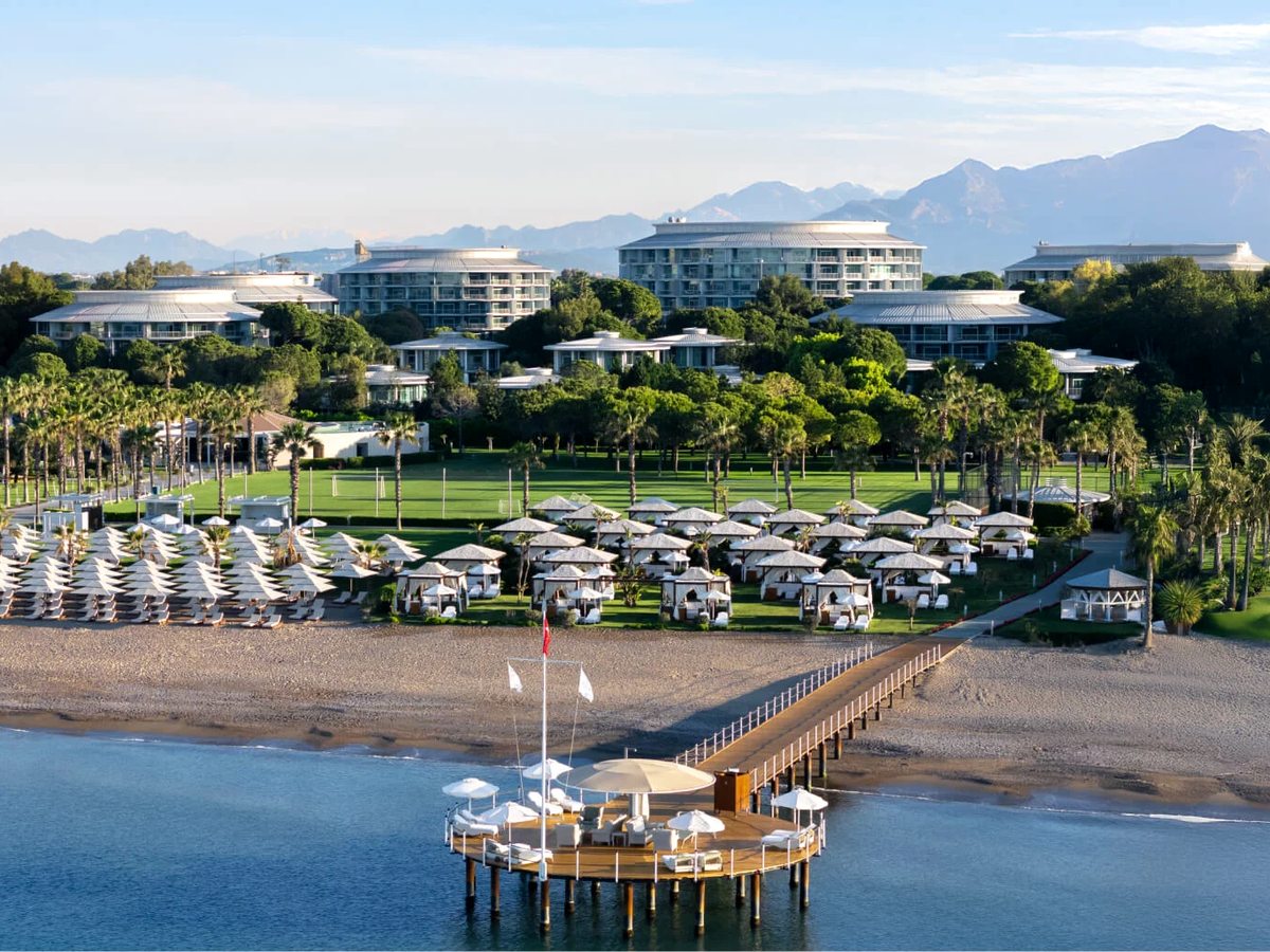 Aerial view of the resort's beachfront, with sunbeds and cabanas along the sand, a pier into the sea and the resort buildings among pines behind.