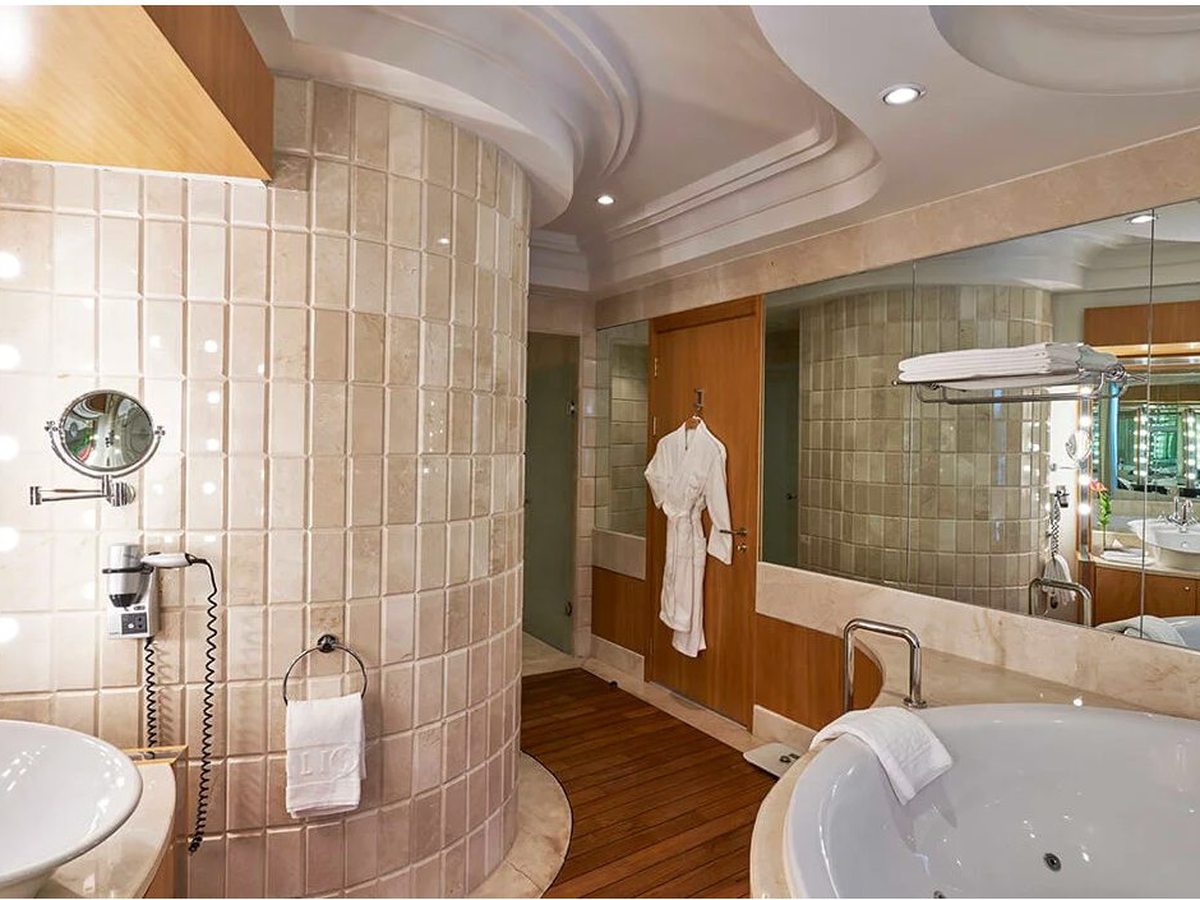A marble-tiled suite bathroom with a corner whirlpool bath, a walk-in shower, twin basins and a bathrobe on the wall.