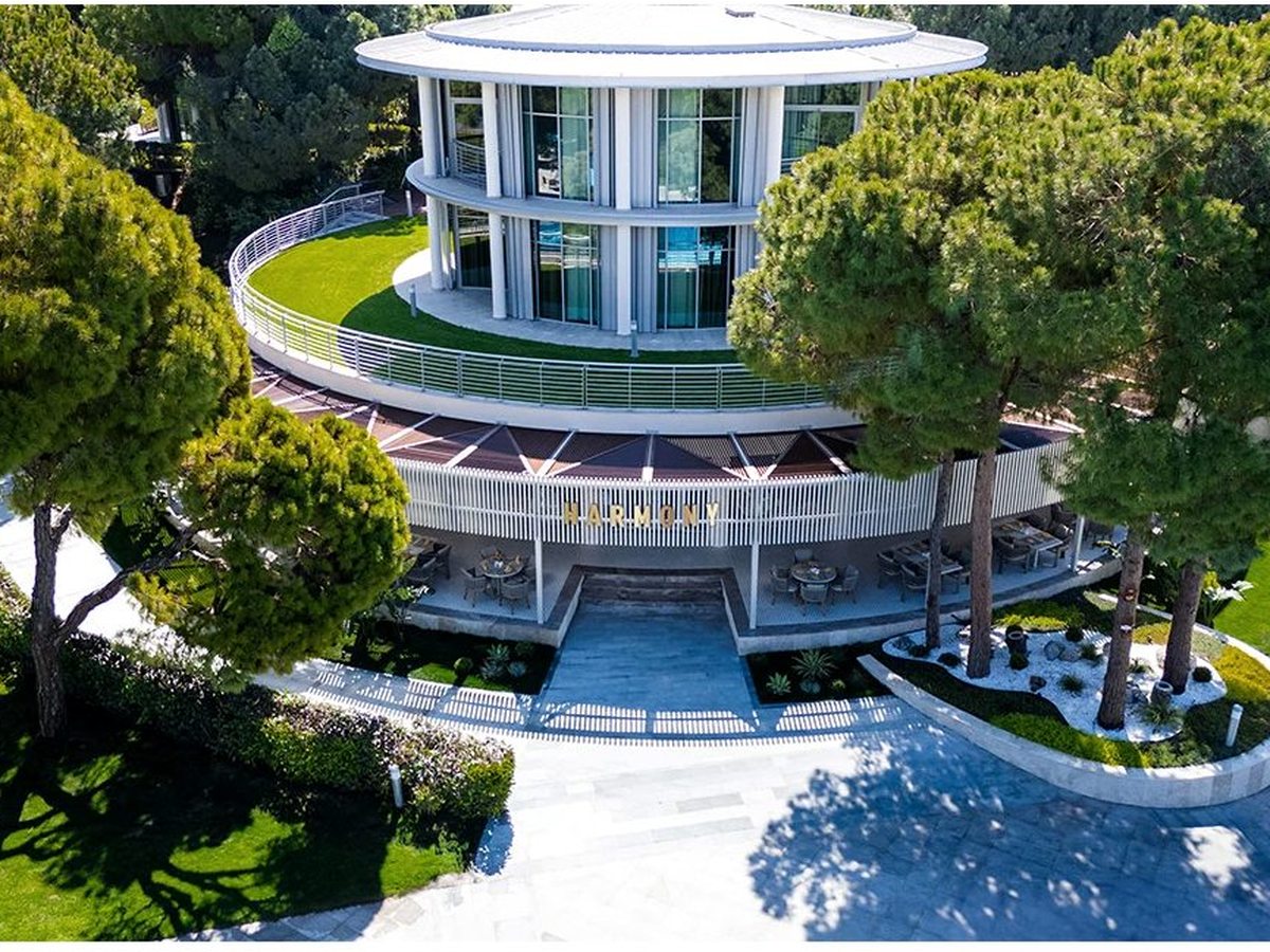 Aerial view of a round white resort building with a wraparound balcony, a grassed rooftop and glass facade, ringed by pine trees and garden paths.