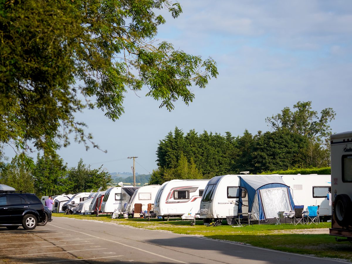 A row of touring caravans with awnings on a grass pitch field, trees behind.