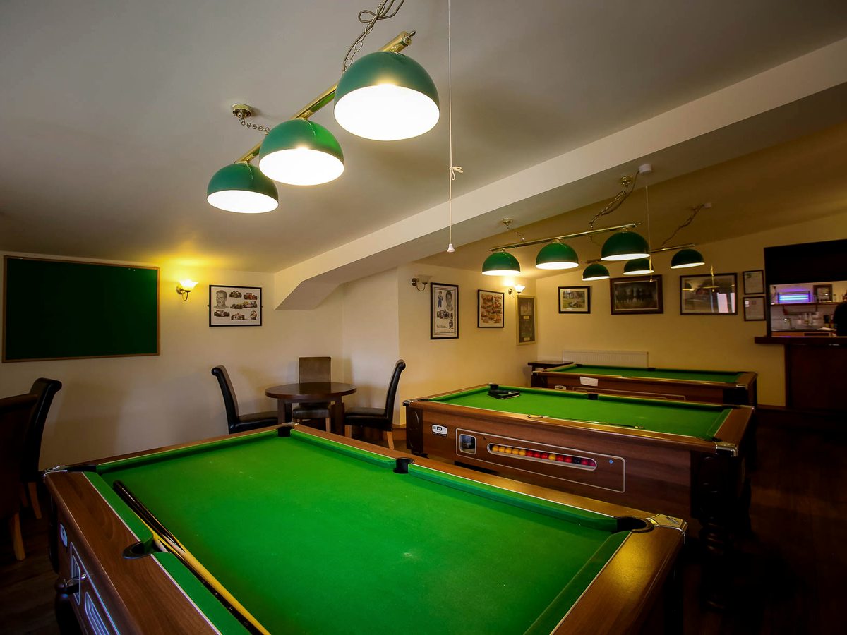 An indoor games room with two full-size pool tables under low green pendant lights and a bar beyond.