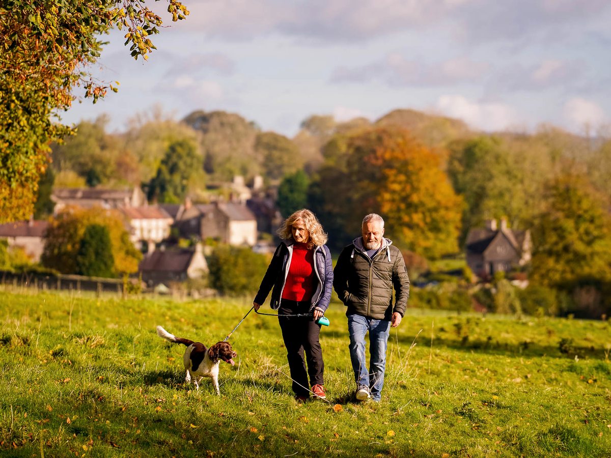 A couple with a dog walking a grassy path with a village and rolling hills beyond in autumn light.