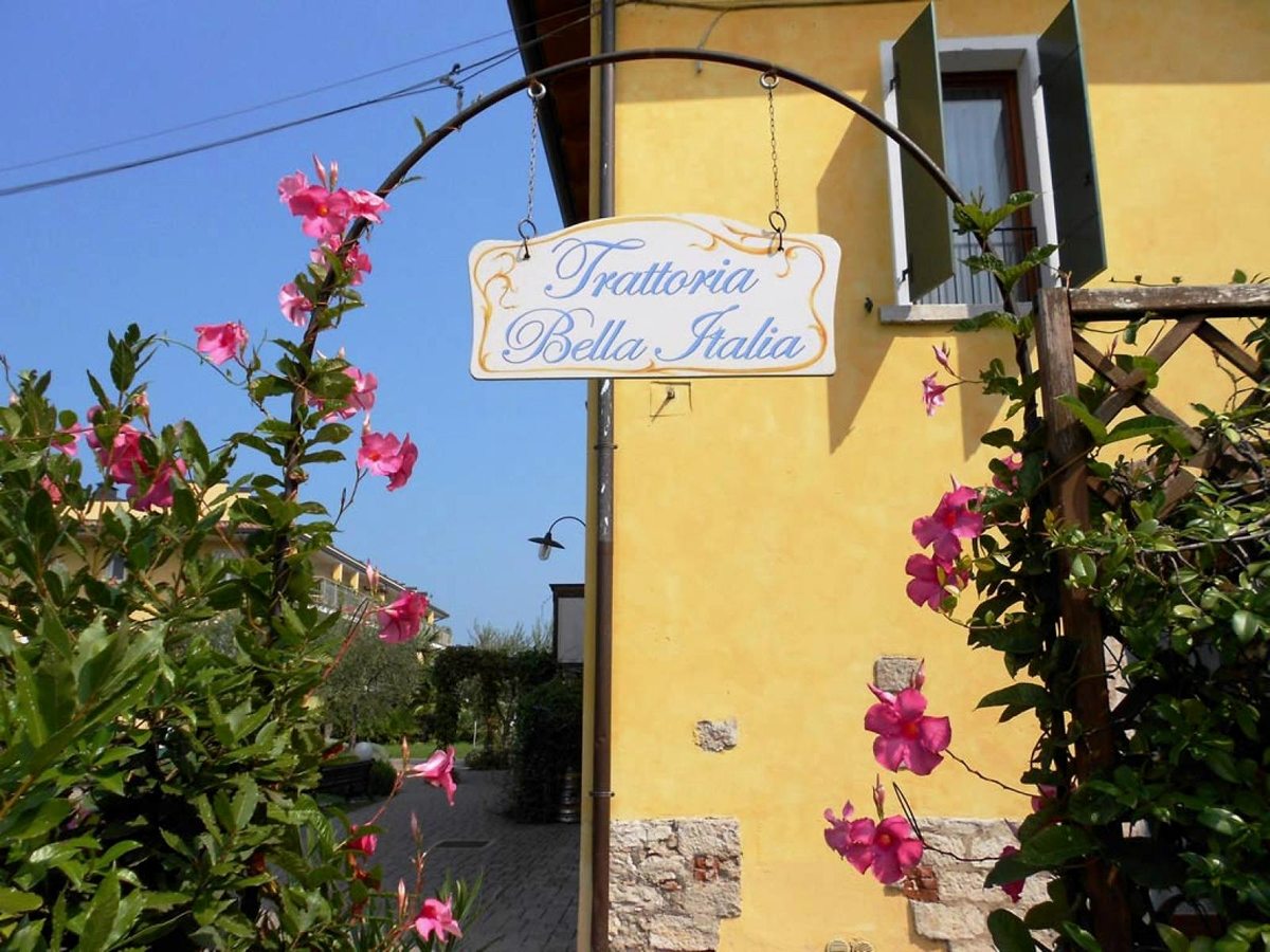 A hanging Trattoria Bella Italia sign on a yellow rendered building, framed by pink flowering vines.