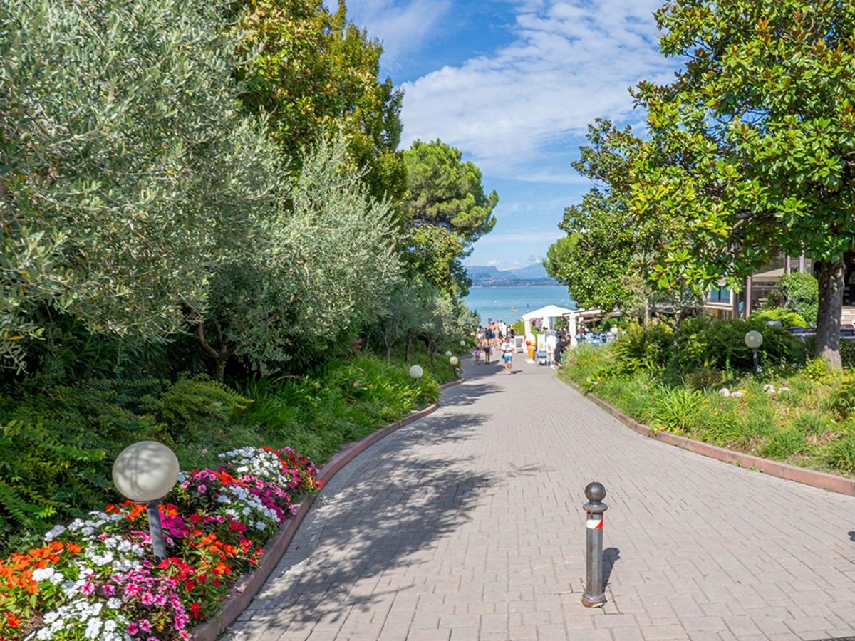 A gently sloping paved path lined with flower beds, mature trees and globe lamp posts, leading down toward the lake.
