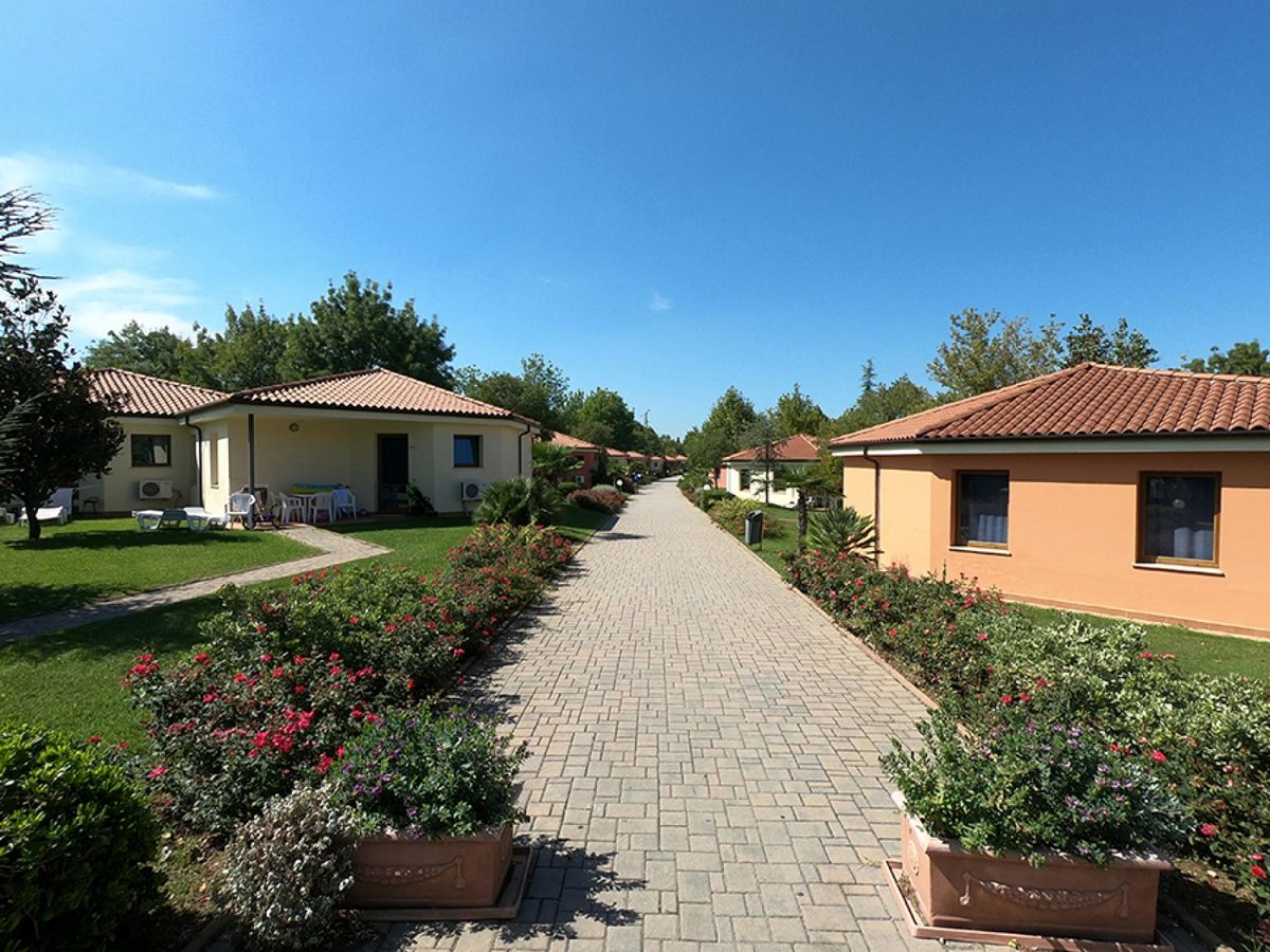 A paved avenue between single-storey bungalows with red-tiled roofs, rose beds and clipped hedges under a clear blue sky.