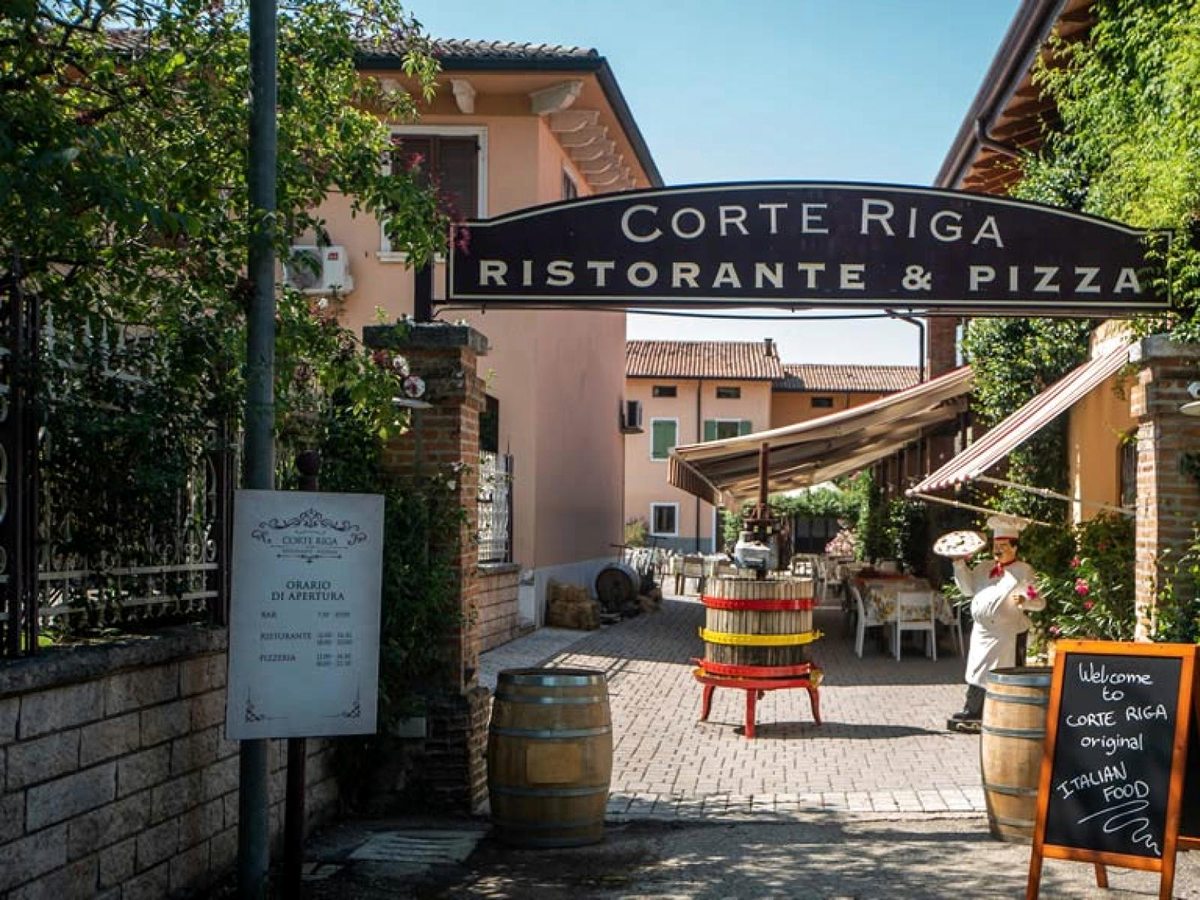 The entrance to Corte Riga Ristorante and Pizza, with an overhead sign, wine barrels and a covered dining courtyard beyond.