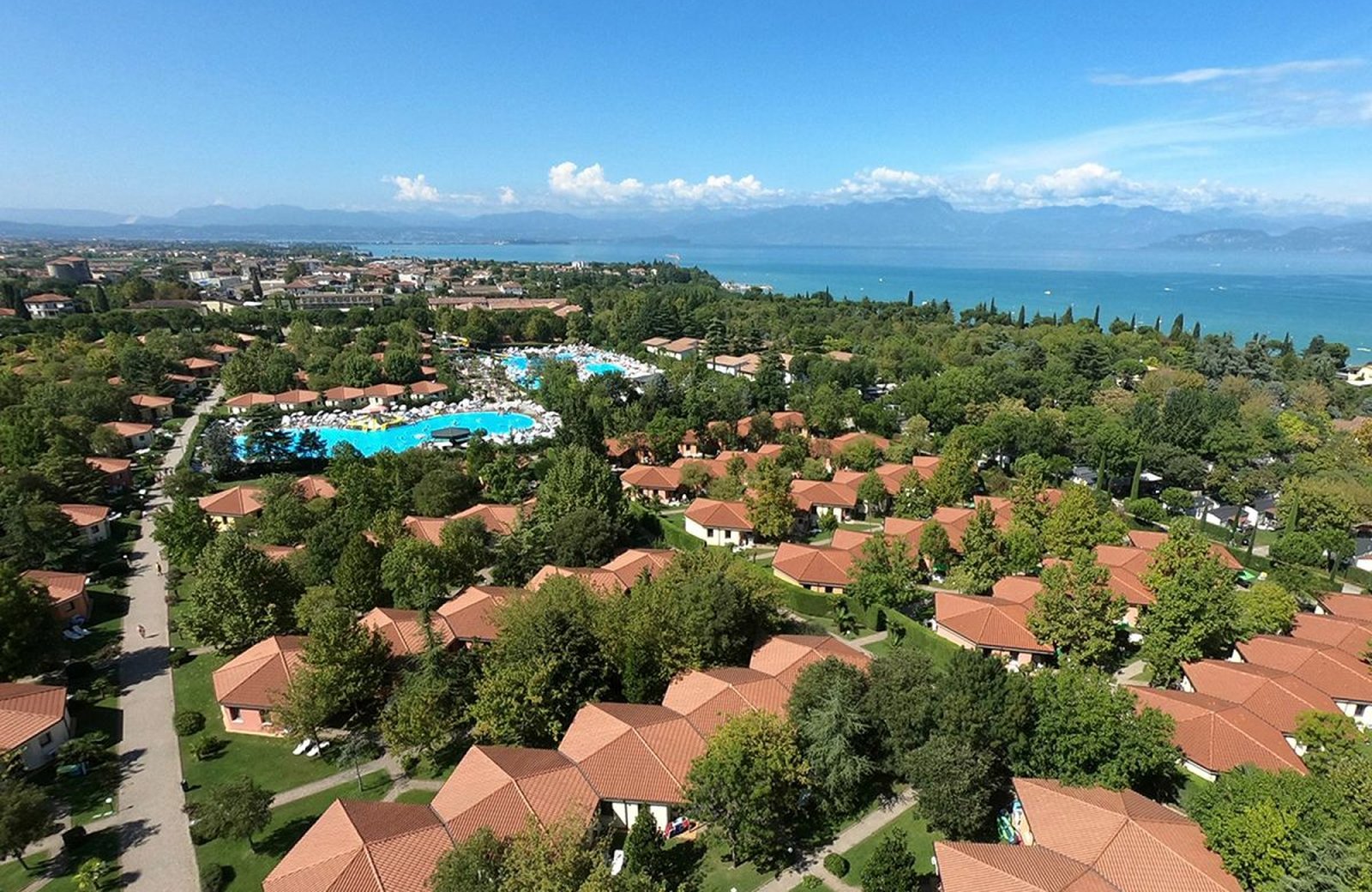 An aerial view of Camping Bella Italia at Peschiera del Garda: red-roofed bungalows among wooded grounds, the aquapark pools and Lake Garda with the mountains beyond.