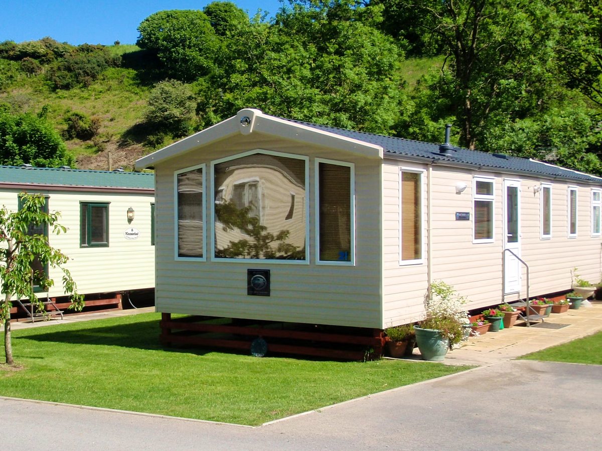 A cream two-tone static caravan with a bay window on a mown grass pitch, steps up to the door and a wooded hillside behind.