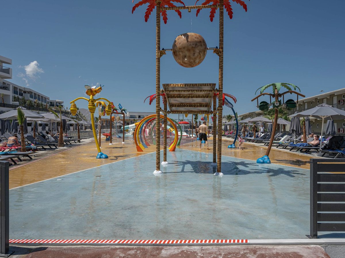 Zero-depth splash pad with palm-shaped spray features, a tipping bucket and rainbow water arches, sun loungers along both sides.