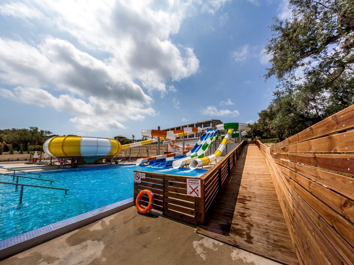 Coloured water slides beside a pool, a wooden ramped walkway leading up to the slide start, a white resort building behind.
