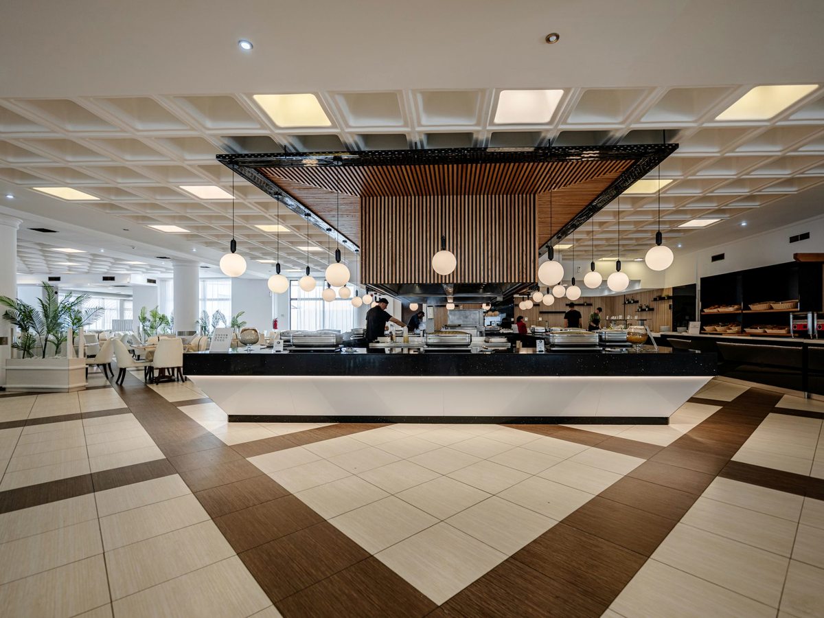 Large indoor buffet restaurant with serving counters under a slatted-wood canopy and pendant lights over a tiled floor.