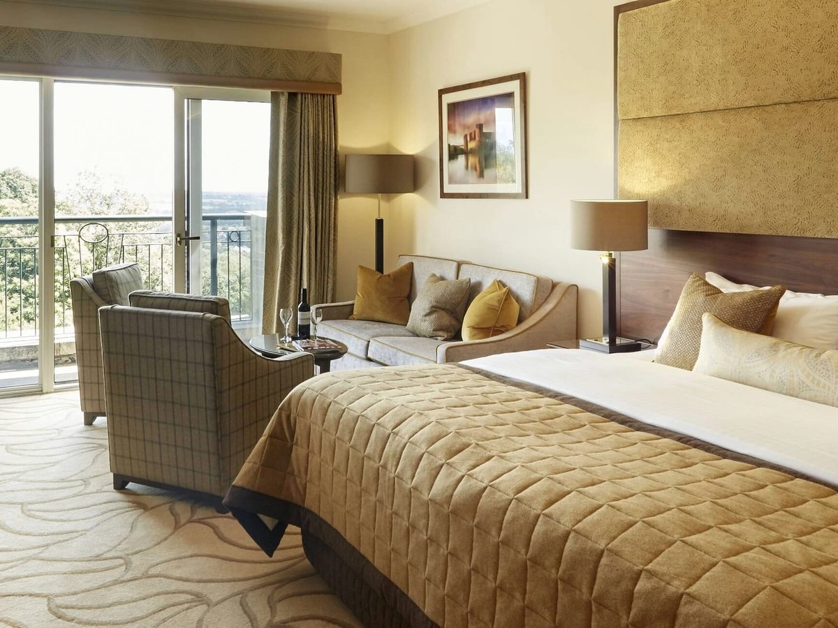 A king-bedded Resort Hotel room with full-height windows and balcony doors onto a leafy view, a desk and an armchair.