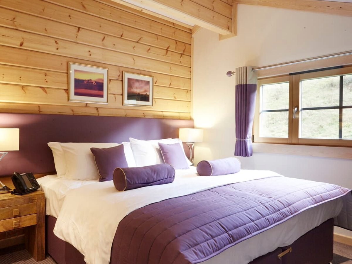 A wood-panelled lodge bedroom with a king bed, a purple runner and bolster cushions, framed prints and a curtained window.