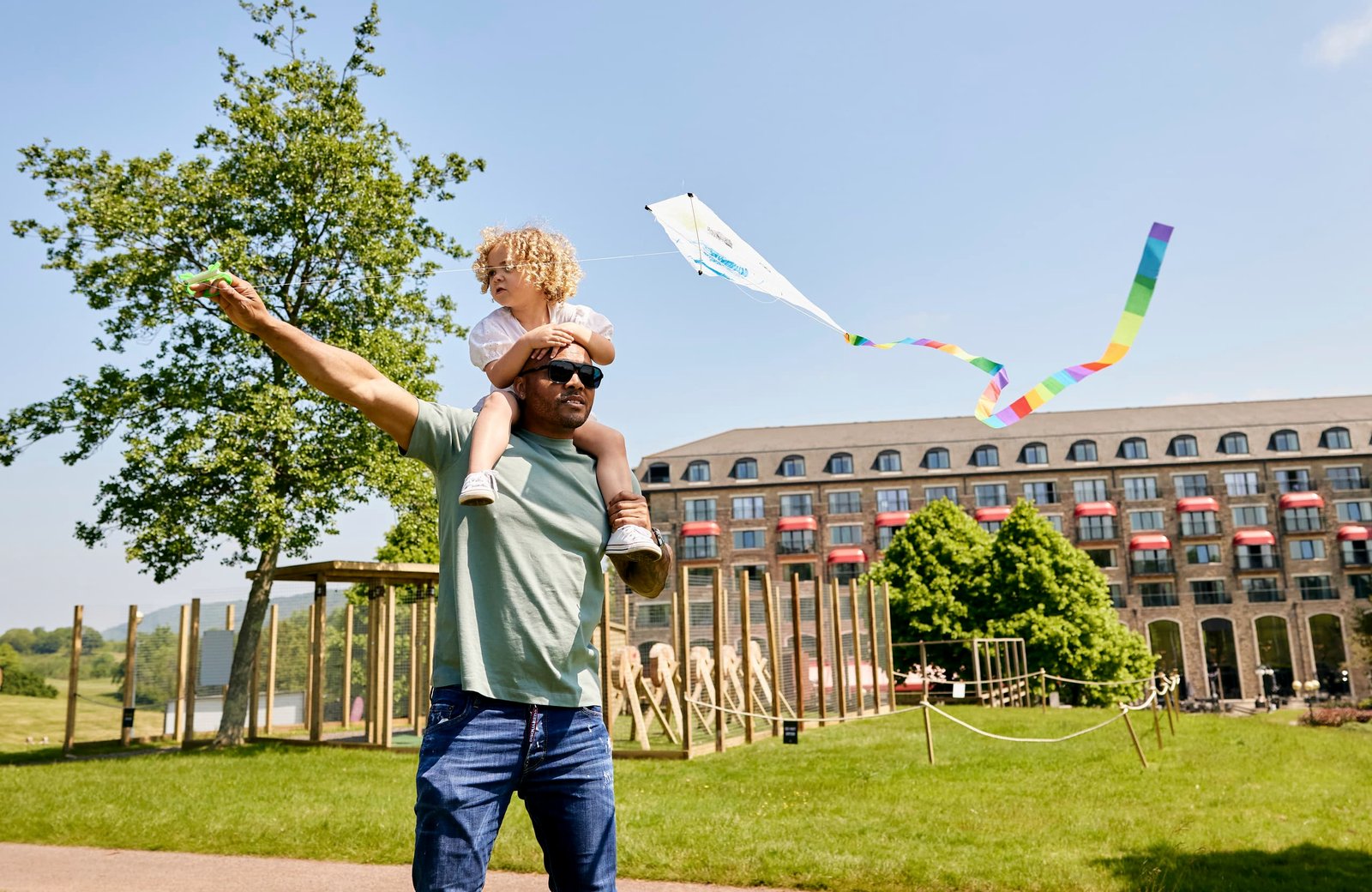 A father with a young child on his shoulders flying a kite on the lawn in front of the Celtic Manor Resort Hotel.