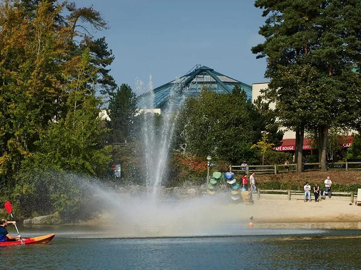 A wide lake with a tall fountain plume, a red kayak on the water and village buildings among trees on the far bank.